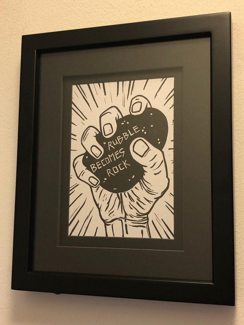 A Lino print made with black ink on white paper, depicting a closeup of a hand clutching a rock with the words, “Rubble becomes Rock” inscribed on it.
