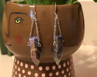 May include: A pair of silver earrings with purple and green stone pendants. The stones are long and pointed, and the earrings are hanging from a chain.