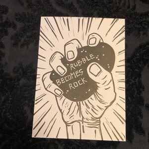 Puede incluir: Una ilustraci&oacute;n en blanco y negro de una mano sosteniendo una roca con el texto "Rubble Becomes Rock" en ella.