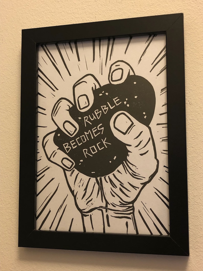 Puede incluir: Ilustraci&oacute;n en blanco y negro de una mano sosteniendo una gran roca con el texto "Rubble Becomes Rock" impreso en ella.