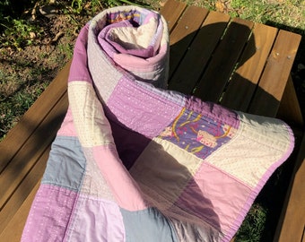 May include: A purple and white patchwork quilt with a floral print on one of the squares. The quilt is rolled up and sitting on a wooden bench.