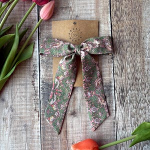 May include: A pink and green floral patterned fabric hair bow tied in a bow shape. The bow is on a brown paper tag with the text "Pretty Little Head" in a circle.