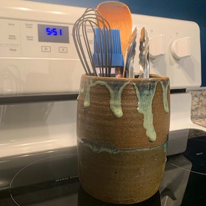May include: A brown ceramic utensil holder with green and blue glaze drips. The holder is filled with kitchen tools, including a wooden spoon, a whisk, a blue silicone basting brush, and a pair of tongs.