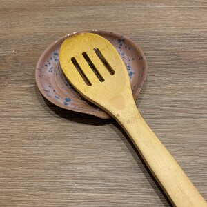 May include: A wooden spoon with a slotted design rests on a pink and blue ceramic spoon rest with a speckled pattern.