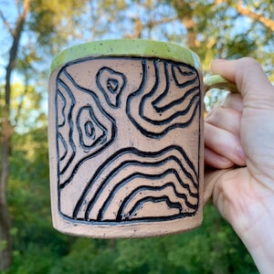 May include: A ceramic mug with a green rim and a brown body. The mug has a black and white design of abstract lines and shapes.