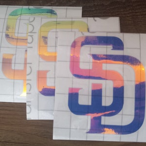 May include: Several iridescent vinyl decals featuring the letters "SD" in a stylized font. The decals have a holographic effect, displaying a range of colors including blue, orange, and yellow. The decals are on a grid-patterned backing.