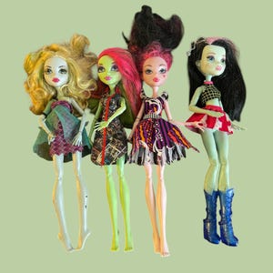 May include: Four Monster High dolls with unique features. One doll has blonde curly hair and a plaid dress. Another has green skin and red hair, wearing a dress with a newspaper print. The third doll has pink hair and a purple dress. The last doll has black hair and a red skirt.