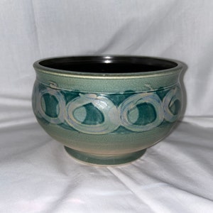 May include: A green ceramic bowl with a white and blue design. The bowl has a rounded shape and a wide rim.