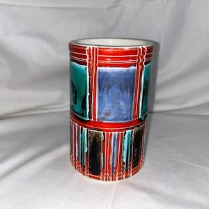 May include: A cylindrical ceramic vase with a geometric pattern in shades of red, blue, green, black, and white.