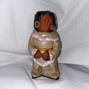 May include: A ceramic figurine of a Native American figure. The figure has dark hair, a brown face, and is wearing a tan outfit with fringe and a yellow belt. The figure has blue earrings.