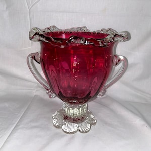 May include: A ruby red glass bowl with clear glass handles and a clear glass base with a scalloped edge. The bowl has a fluted design.