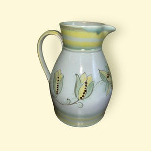 May include: A ceramic pitcher with a handle and spout. The pitcher is primarily light blue with yellow and green floral designs. The top rim features horizontal yellow stripes. The pitcher is a decorative item.