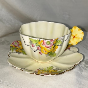 May include: A vintage teacup and saucer set with a floral design. The cup has a yellow, pink, and green floral pattern and a yellow flower handle. The saucer has a scalloped edge and a matching floral design.