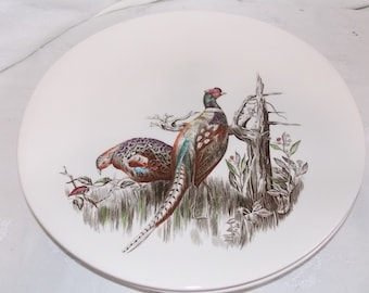 May include: A white ceramic plate featuring a brown and green design of two pheasants on a branch. The pheasants are brown, green, and red.