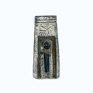 May include: A rectangular ceramic pendant with a textured silver-grey finish. The surface features a central arched motif outlined in blue and brown, containing a dark blue circular element above a vertical blue line, with etched markings at the top.