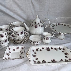 May include: A white tea set with purple floral designs. The set includes a teapot, creamer, sugar bowl, cake stand, and six teacups and saucers.