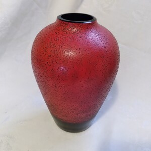 May include: A red ceramic vase with a textured surface and a black rim. The vase is round and has a wide opening.