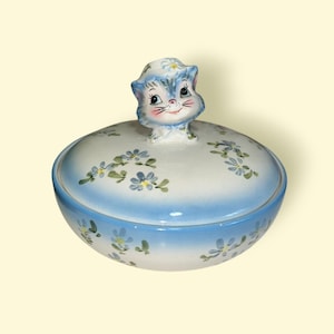 May include: A vintage ceramic trinket dish with a blue and white floral design. The lid features a charming cat figurine. The dish has a light blue base and is decorated with hand-painted blue flowers and green leaves.
