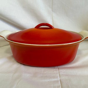 May include: Orange enamel cast iron Dutch oven with a white lid and white handles. The oven is oval shaped.