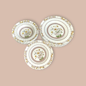 May include: A set of white ceramic plates and bowls with a floral design. The plates and bowls have a scalloped edge and a central floral motif with yellow flowers and green leaves. The plates are stacked in a neat pile.