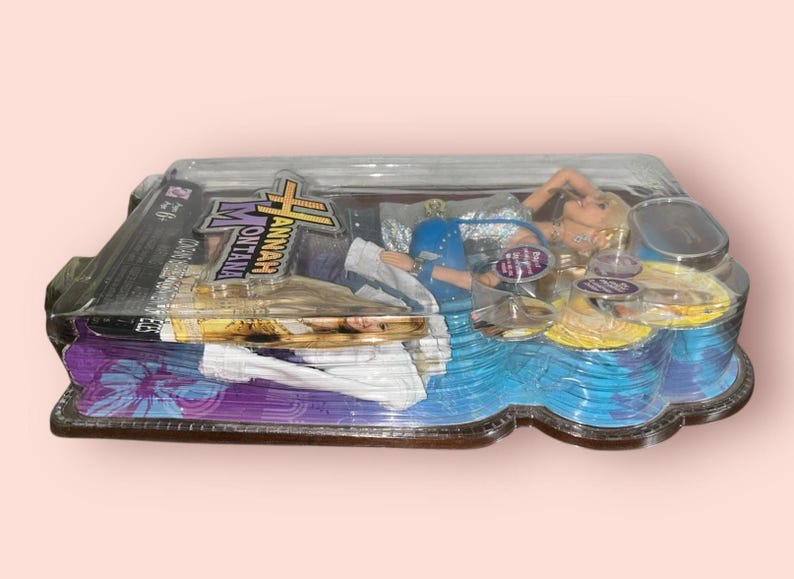 Pu&ograve; includere: Una bambola di Hannah Montana confezionata in una custodia di plastica trasparente. La bambola &egrave; bionda e indossa un abito blu. La confezione presenta il logo di Hannah Montana e altro testo. La custodia ha un design blu e viola.