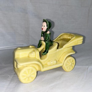 May include: A yellow ceramic figurine of a vintage car with a green-clad figure driving. The car has a removable top and is designed as a planter.