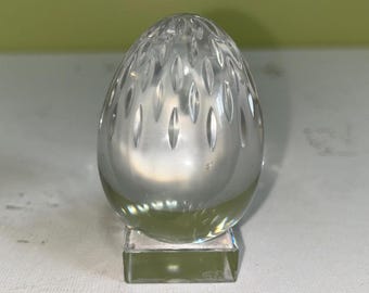 May include: A clear, egg-shaped crystal sculpture with a textured surface. The egg sits on a small, square, clear base. The egg has a series of vertical, oval-shaped cuts that run from top to bottom.