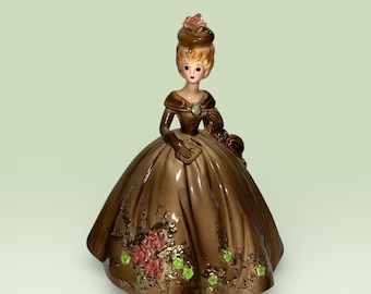 May include: A vintage porcelain figurine of a woman in a long brown dress with a full skirt. The dress is decorated with pink roses and green accents. The woman has blonde hair and is wearing a matching hat.