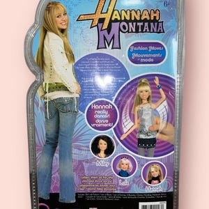 Pu&ograve; includere: Una bambola di Hannah Montana in una confezione di plastica trasparente. La confezione presenta immagini di Hannah Montana, Miley e Lola. Il testo sulla confezione recita "Fashion Moves" e "Hannah really dances!"
