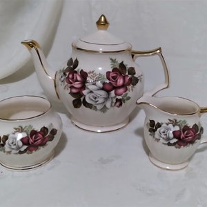 May include: A white teapot, sugar bowl, and creamer with gold trim and a floral design featuring red and white roses.