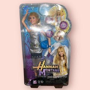 Pu&ograve; includere: Una bambola di Hannah Montana nella sua confezione originale. La bambola indossa un vestito argentato e tiene in mano una borsetta blu. La confezione include il logo di Hannah Montana e il testo "CONTAINS 16 PIECES".