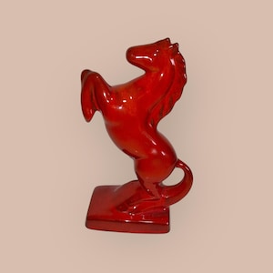 May include: A glossy, red ceramic horse figurine rearing up on its hind legs. The sculpture is mounted on a rectangular base. The horse has a flowing mane and tail, and the overall design is stylized and elegant.
