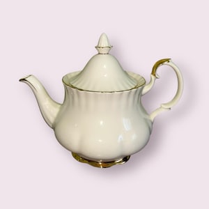 May include: A cream-coloured porcelain teapot with gold trim and handle. The teapot has a rounded body, a curved spout, and a lid with a finial. The base of the teapot is also trimmed in gold. A classic piece for serving tea.