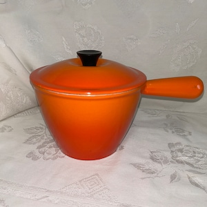May include: Orange enamelled cast iron pot with a black knob on the lid and a handle. The pot is a gradient colour from a darker orange to a lighter orange.