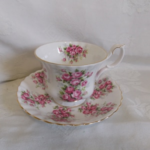 May include: A white teacup and saucer with a pink rose floral pattern. The cup has a gold rim and the saucer has a scalloped edge.