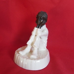 May include: A white porcelain figurine of a young person sitting with their chin resting on their hand. The figurine has a white base and the person's head and hands are painted brown.