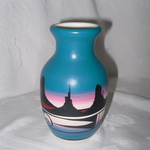 May include: A turquoise ceramic vase with a painted scene of a desert landscape with a pink and purple sunset. The vase has a white rim and a white band around the middle.