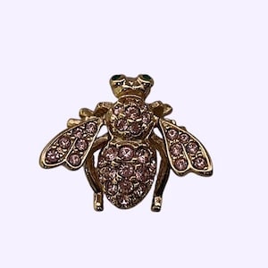 May include: A gold-coloured bee brooch with pink crystal detailing. The bee's body and wings are embellished with sparkling pink crystals, and it features green crystal eyes. This brooch is a decorative accessory.