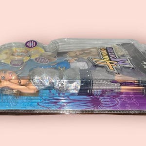 Pu&ograve; includere: Una bambola di Hannah Montana in una confezione di plastica trasparente. La bambola ha i capelli biondi e indossa un top argentato e una gonna nera. La confezione presenta il logo di Hannah Montana e altre grafiche.