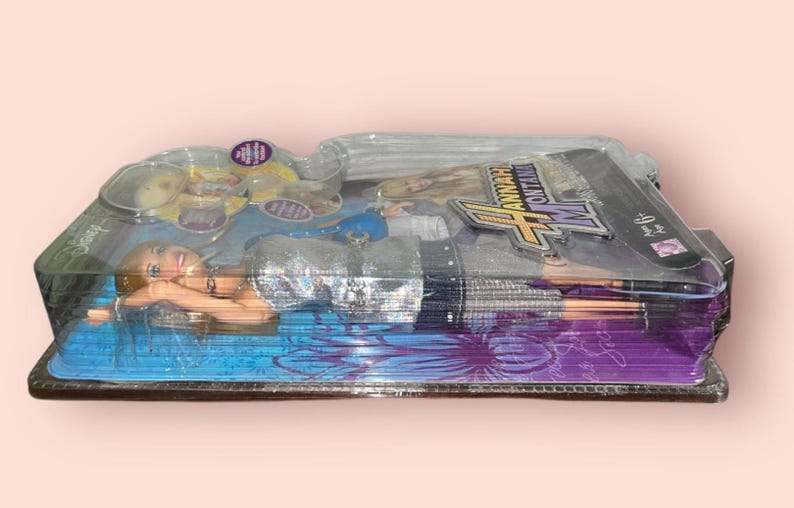 Pu&ograve; includere: Una bambola di Hannah Montana in una confezione di plastica trasparente. La bambola ha i capelli biondi e indossa un top argentato e una gonna nera. La confezione presenta il logo di Hannah Montana e altre grafiche.