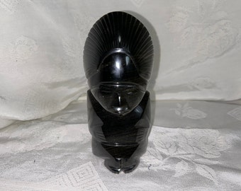 May include: Black obsidian sculpture of a person with a headdress. The sculpture is carved in a stylized, abstract style.