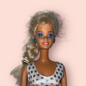 May include: A vintage doll with blonde hair styled in a side ponytail. The doll has blue eye shadow and matching blue earrings. She wears a sleeveless silver dress with blue star patterns. The doll is set against a light pink background.