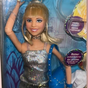 Pu&ograve; includere: Una bambola Disney con capelli biondi, che indossa un top argentato con paillettes, una borsa blu e una collana a girocollo. La bambola &egrave; in una confezione di plastica trasparente con il testo "really dancer! danse raiment!" e "Bonus jacket & purse!"