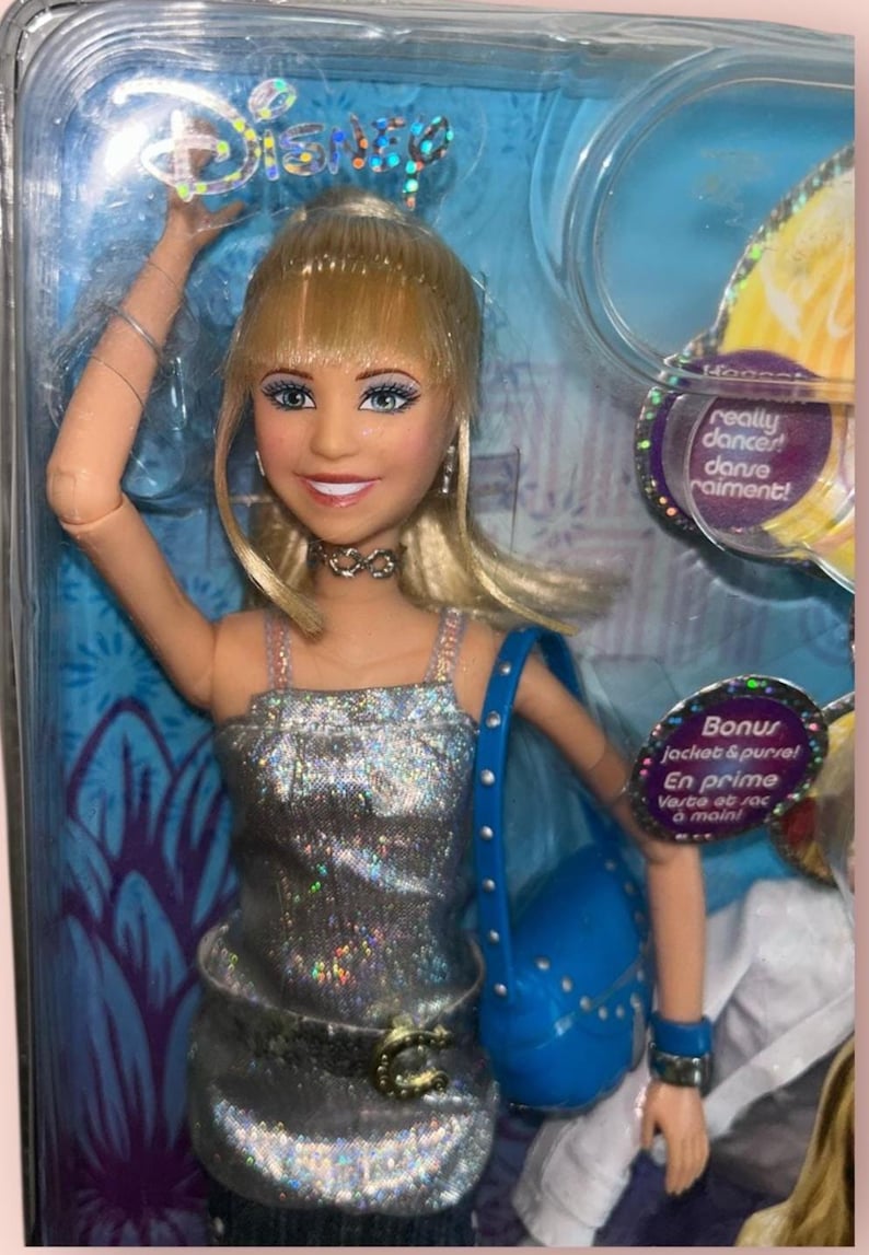 Pu&ograve; includere: Una bambola Disney con capelli biondi, che indossa un top argentato con paillettes, una borsa blu e una collana a girocollo. La bambola &egrave; in una confezione di plastica trasparente con il testo "really dancer! danse raiment!" e "Bonus jacket & purse!"