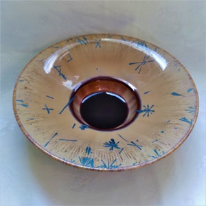 May include: A round, shallow glass bowl with a brown and purple iridescent glaze. The bowl has a decorative design of blue lines and stars.