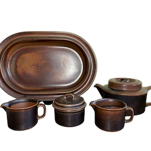 May include: A brown stoneware tea set comprising a teapot, creamer, sugar bowl, and oval serving platter. The set has a rustic, earthy appearance.