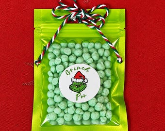 Puede incluir: Una bolsa resellable de color verde lima llena de pequeños dulces esféricos de color verde claro. La bolsa está atada en la parte superior con un cordel trenzado rojo, blanco y verde. Una pegatina blanca circular al frente muestra un personaje verde y el texto Grinch Poo.
