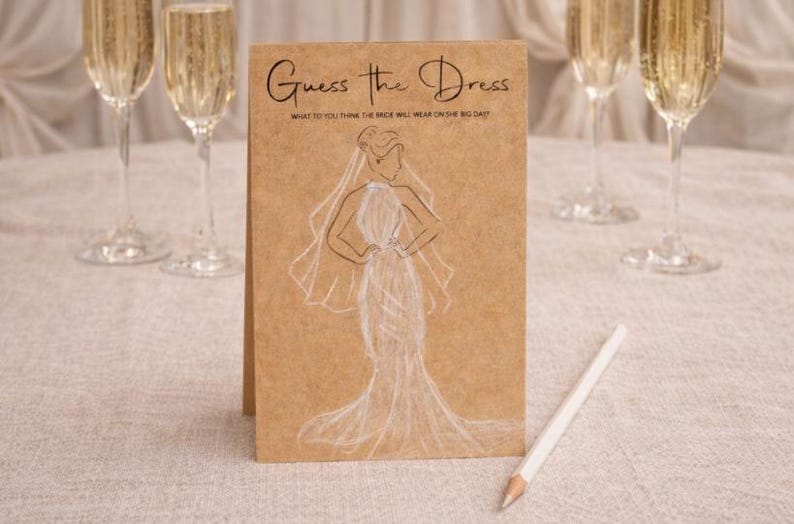 May include: A kraft paper card with the text "Guess the Dress" and a sketch of a wedding dress. A white pencil rests on the table. Champagne flutes filled with a golden liquid are in the background. The card is for a bridal shower game.