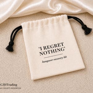 May include: A cream-colored fabric drawstring pouch featuring black text that reads 'I REGRET NOTHING' followed by the words 'hangover recovery kit' in a smaller font. The pouch has black cord drawstrings with knotted ends on both sides.