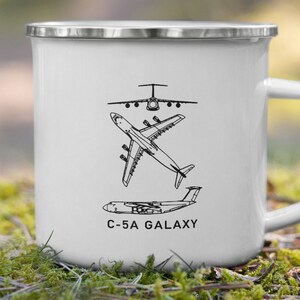 Könnte beinhalten: Weißer Emaille-Becher mit silbernem Rand und Henkel. Der Becher zeigt schwarze Strichzeichnungen von drei verschiedenen Flugzeugen, darunter eine C-5A Galaxy. Der Text "C-5A GALAXY" ist darunter gedruckt.